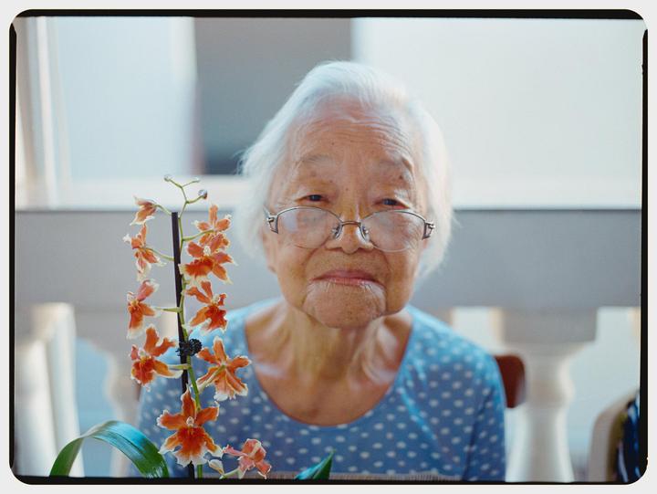 A woman with white hair and glasses looks at the viewer, with an orchid plant in the foreground.