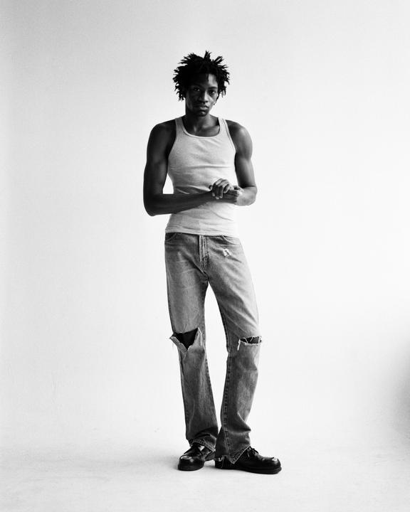 A man with dreadlocks wearing a tank top and ripped jeans stands against a white background.