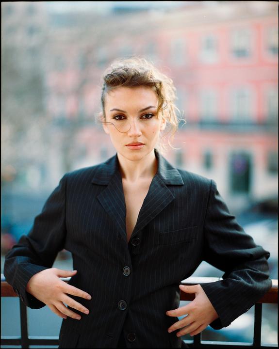 A woman in a pinstripe blazer stands on a balcony with a blurred building in the background.