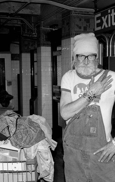 A man with a long white beard and glasses stands in a subway station next to a shopping cart filled with clothes.