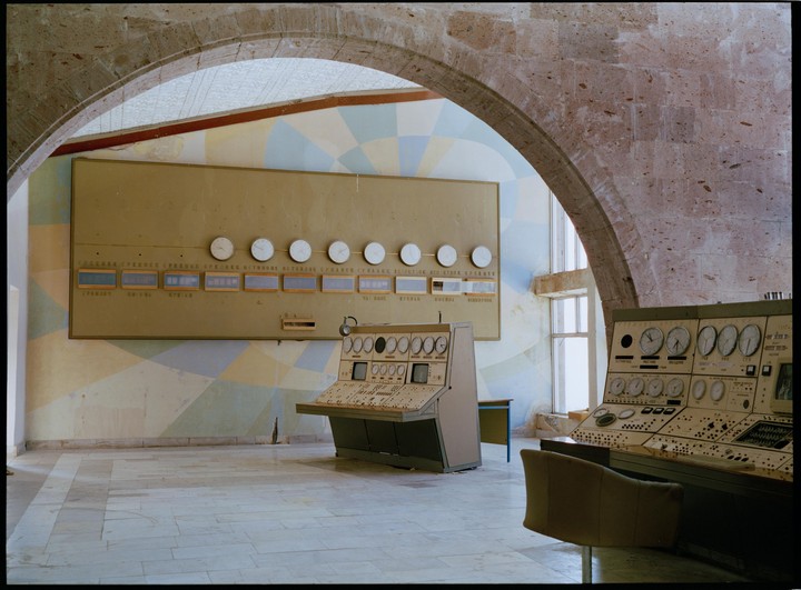 Two large control panels with numerous dials and buttons sit in a room with a large clock display on the wall and an arched stone ceiling.