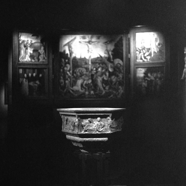 A stone baptismal font with carved figures is illuminated in a dark room with paintings on the wall.