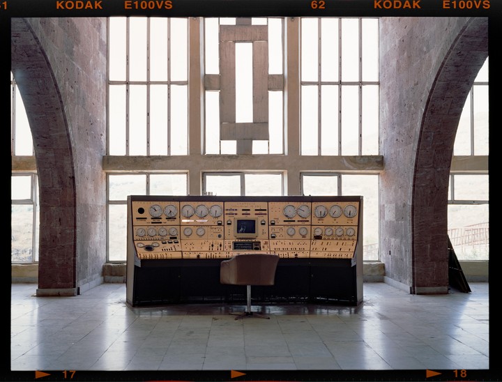 A large control panel with numerous dials and buttons sits in a spacious room with arched walls and tall windows.
