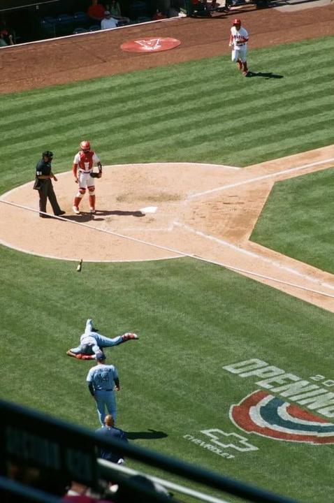 A baseball player lies on the grass field while other players and an umpire stand on the dirt infield.
