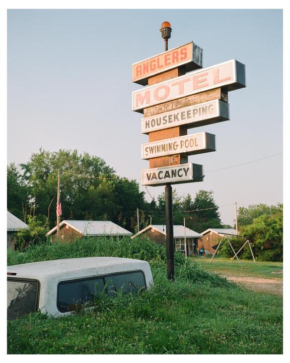 A tall, weathered motel sign stands amidst overgrown grass, with motel buildings and a truck cap visible in the background.