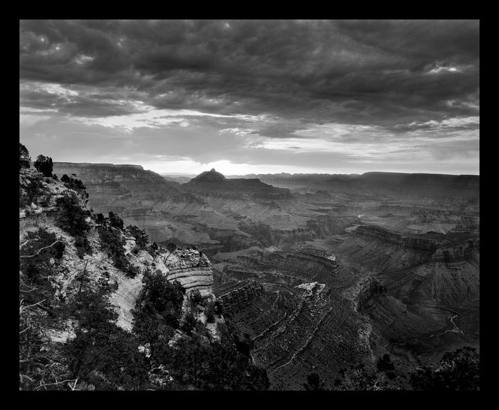 The Grand Canyon is visible under a cloudy sky with the sun rising over the horizon.
