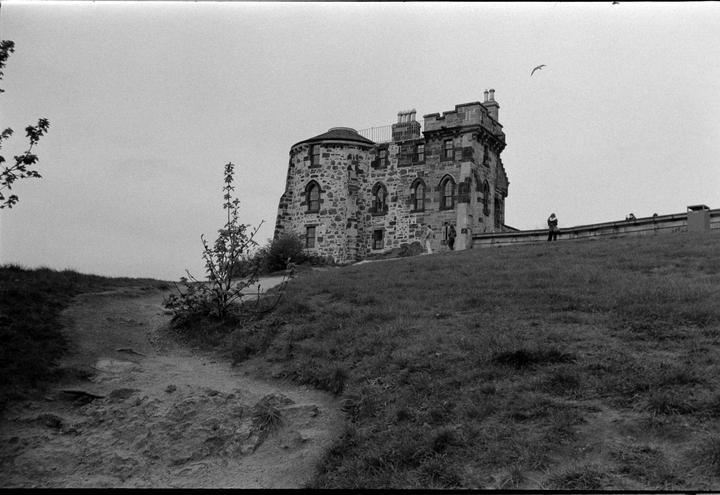 A stone building with turrets stands on a grassy hill with a dirt path in the foreground.