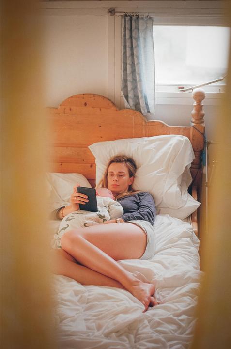 A woman lies in bed with a baby on her chest while looking at an e-reader.