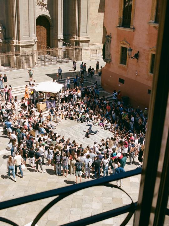 A crowd of people watches a breakdancer perform in a plaza in front of a large building.