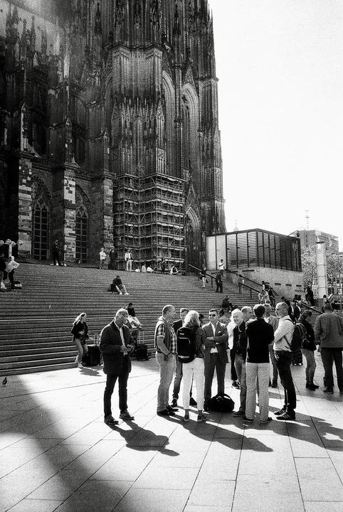 A large group of people are gathered on a paved area in front of a grand cathedral with scaffolding on its facade.
