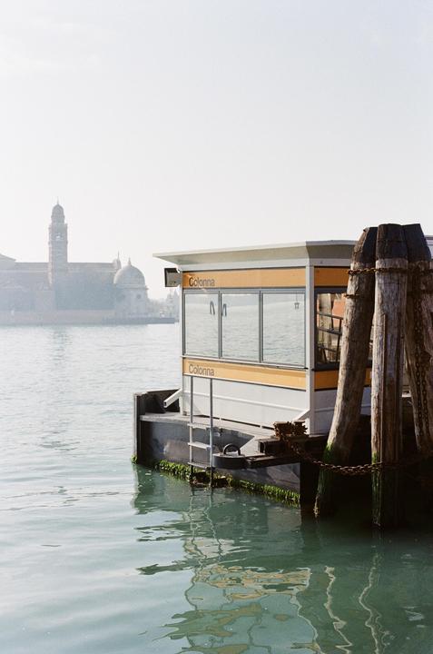 A small ferry terminal building with "Colonna" written on it floats on the water, with a distant city skyline visible in the hazy background.