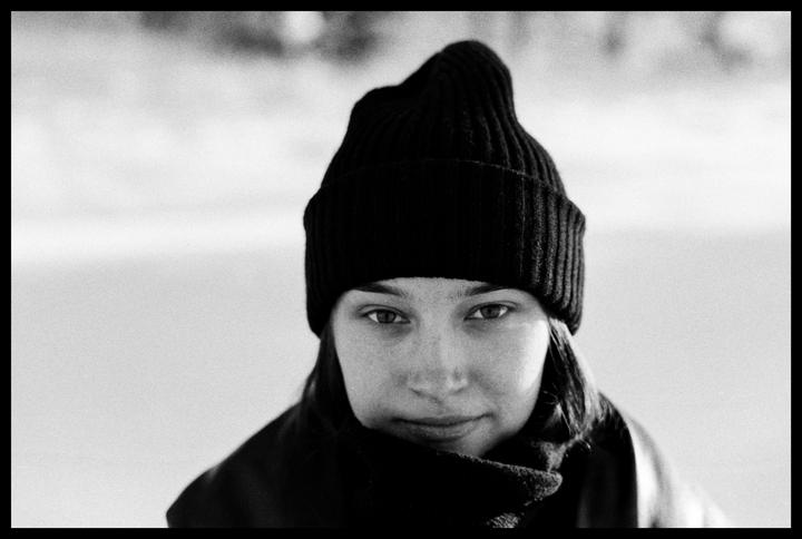A woman wearing a beanie and scarf looks directly at the viewer.