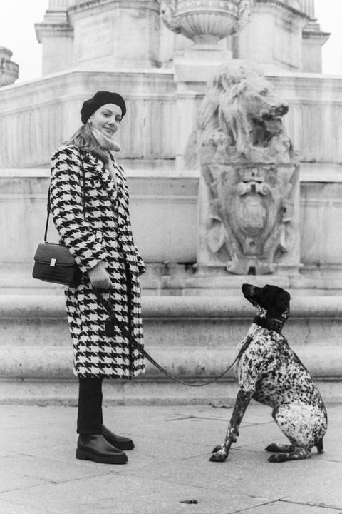 A woman in a houndstooth coat and beret stands next to a spotted dog on a leash, with a stone structure featuring a lion sculpture in the background.