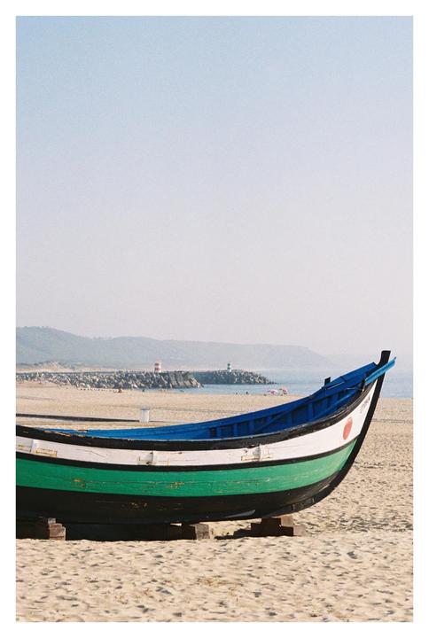 A colorful boat rests on a sandy beach with a distant coastline and lighthouses under a clear sky.