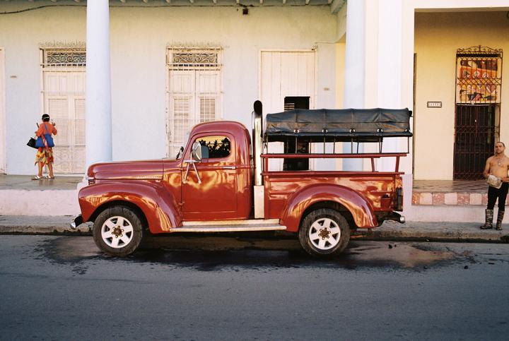 A red pickup truck is parked on a street in front of buildings, with a woman and a man nearby.