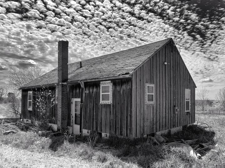 An old wooden house with a brick chimney stands under a sky filled with popcorn clouds.