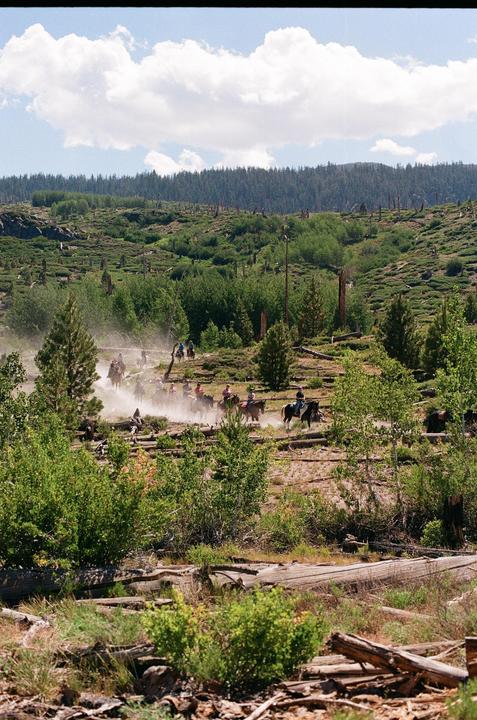A group of people ride horses through a dusty, forested landscape under a cloudy sky.