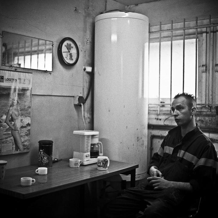 A man sits at a table with coffee cups and a coffee maker in a room with a calendar and a water heater.