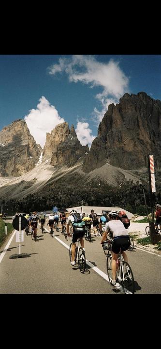 A group of cyclists ride on a road with towering mountains in the background under a blue sky with white clouds.