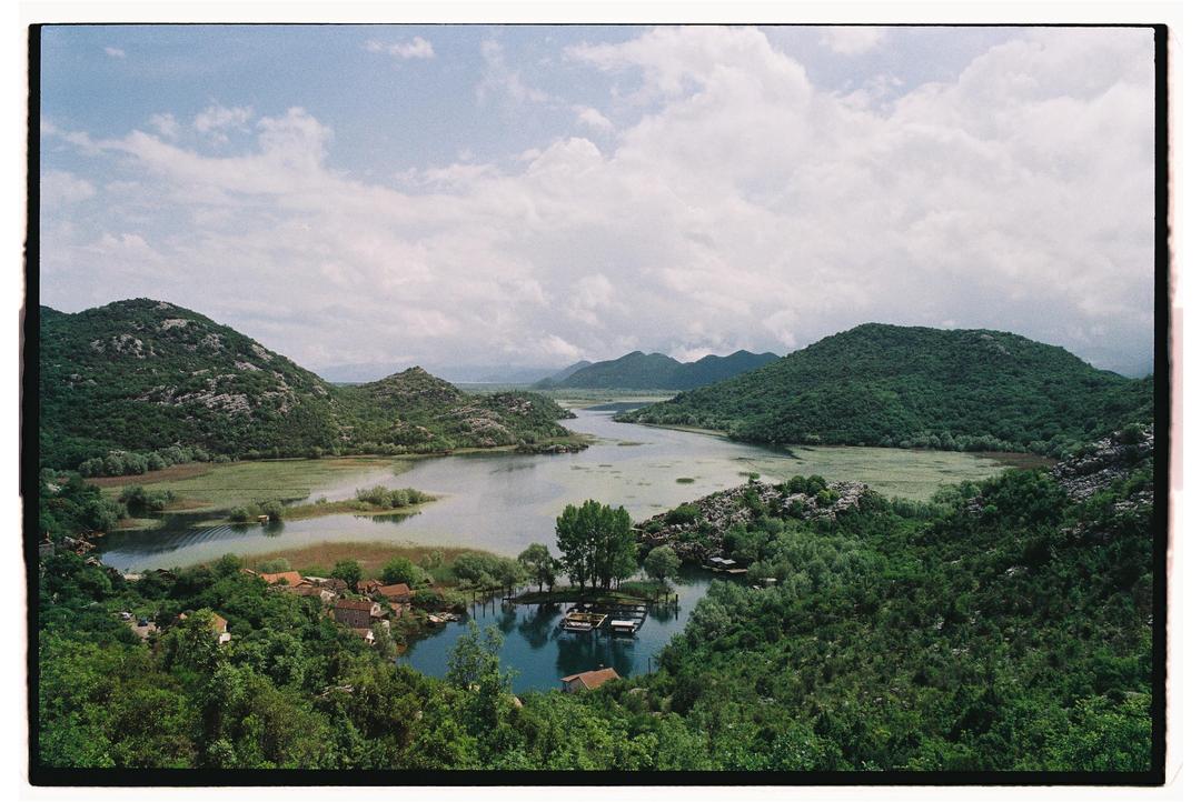 A wide shot shows a lake surrounded by mountains and a small village with houses and boats.