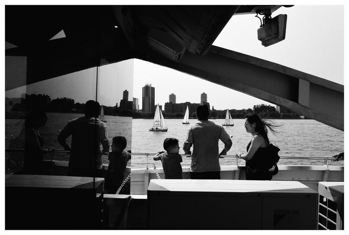 A group of people on a ferry look out at sailboats on the water with a city skyline in the distance.