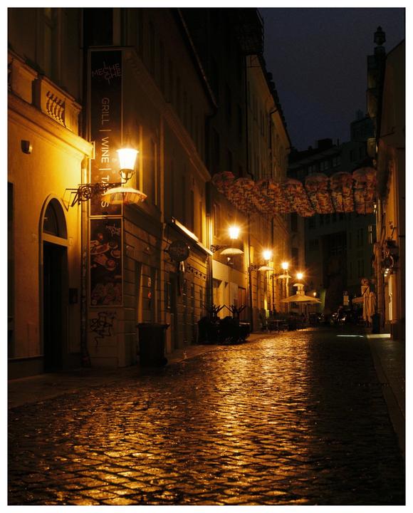 A narrow cobblestone street is illuminated by warm streetlights at night, with reflections visible on the wet ground.