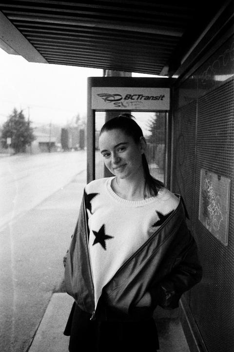 A young woman wearing a star-patterned sweater and a jacket stands at a bus stop.