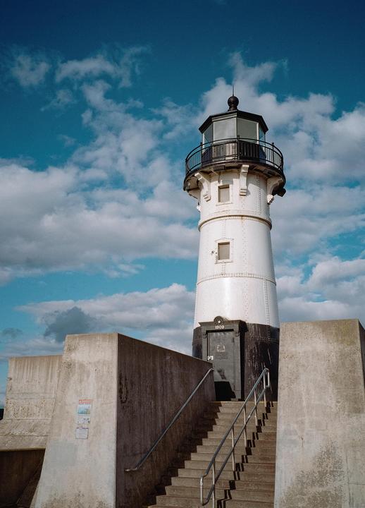 A white lighthouse with a black base stands tall against a blue sky with scattered clouds, with concrete walls and stairs leading up to its entrance.