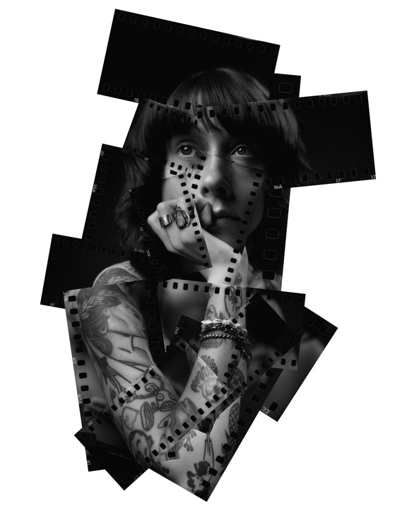 A woman with tattoos on her arms looks up, her face and arms covered by strips of film negatives.
