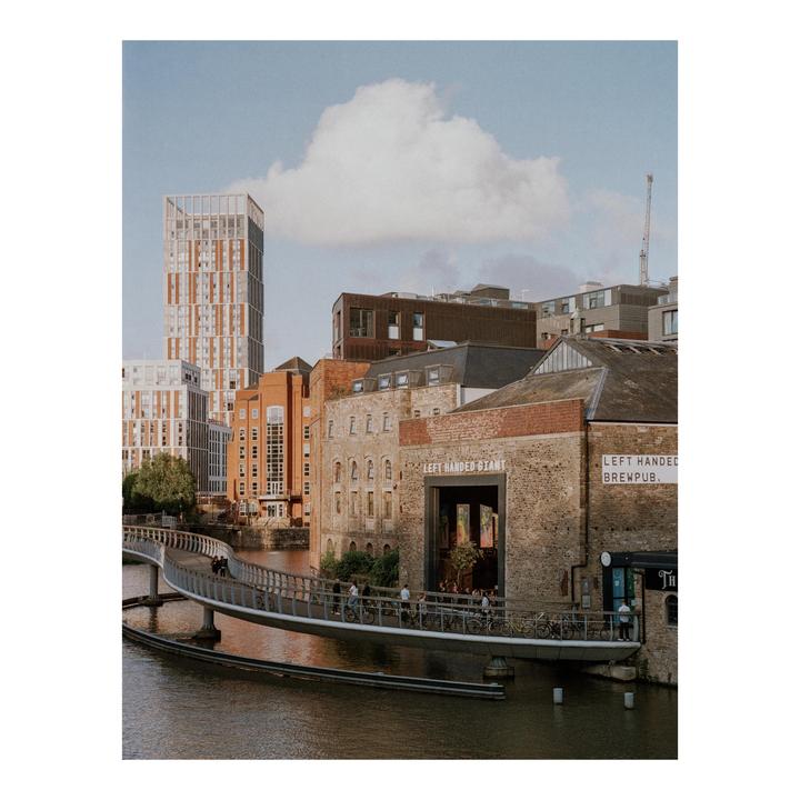 A modern pedestrian bridge crosses a river in a city with a mix of old and new buildings under a partly cloudy sky.
