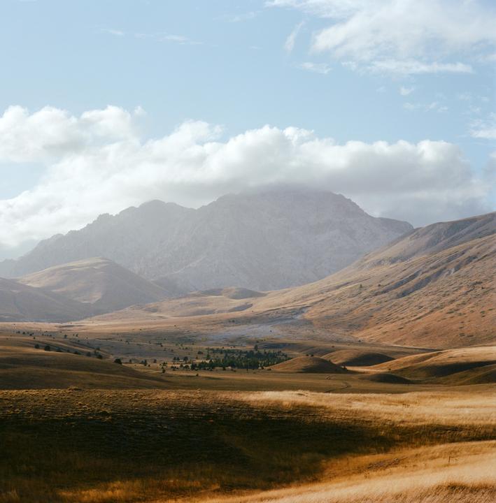 A vast landscape shows rolling hills of dry grass in the foreground and middle ground, with large mountains under a cloudy sky in the background.