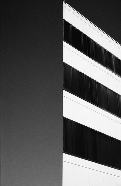 A black and white photograph shows the side of a building with horizontal rows of dark windows and a large, dark, blank wall.
