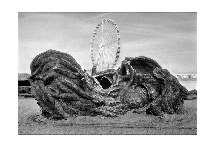 A large sculpture of a head emerging from the ground is in the foreground with a ferris wheel in the background.