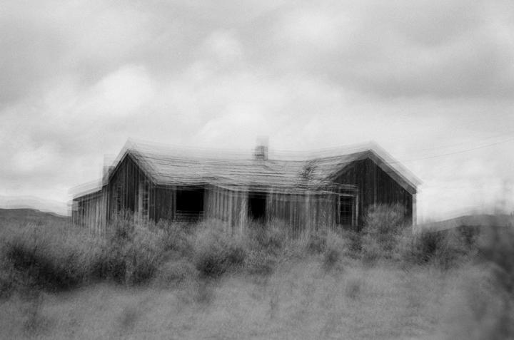 An abandoned wooden house with a chimney is surrounded by tall grass under a cloudy sky.