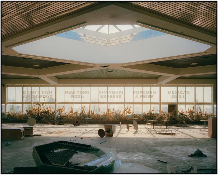 An abandoned food court with restaurant names on the windows is illuminated by sunlight from a large skylight.