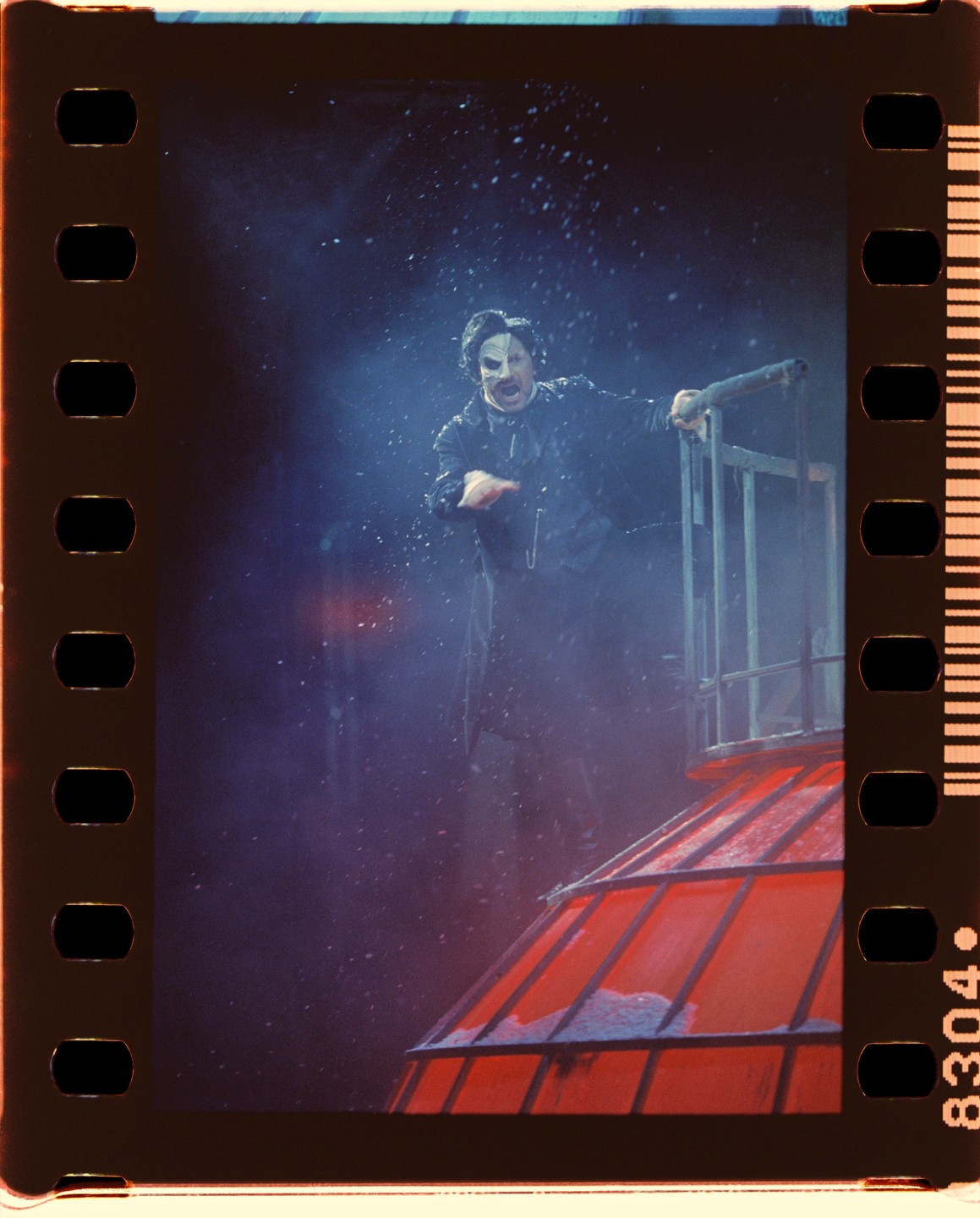 A man in a phantom costume stands on a red roof in the snow.