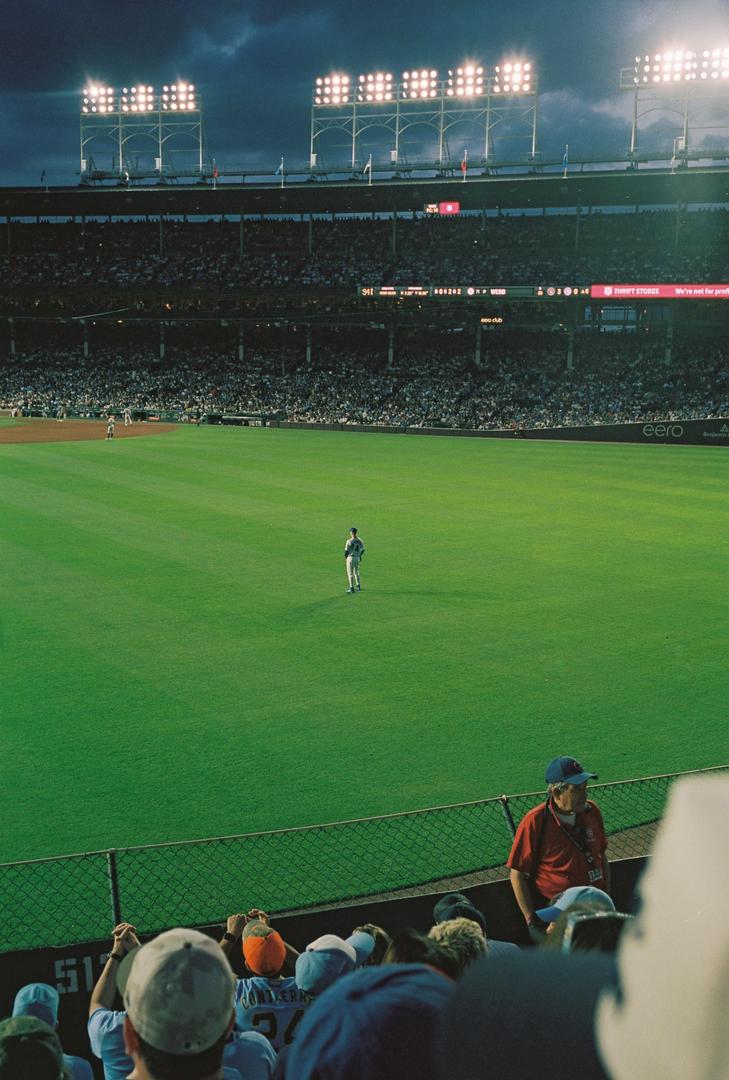 A baseball player stands alone on a green field in a stadium filled with spectators under bright lights at dusk.