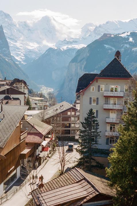 A village with buildings and trees is nestled in a valley surrounded by snow-capped mountains.