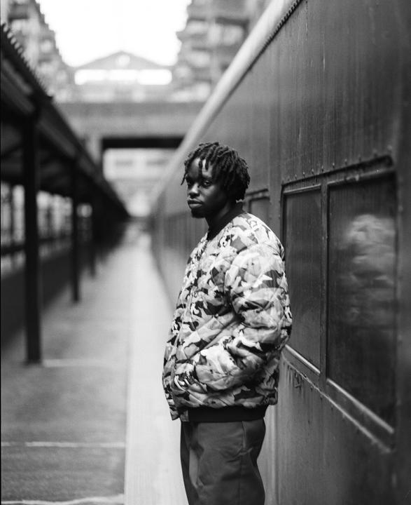 A man with dreadlocks stands on a train platform next to a train car.