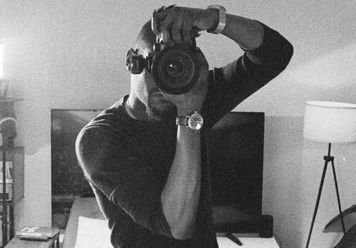 A man holds a camera to his face, taking a self-portrait in a room with a television and a lamp.