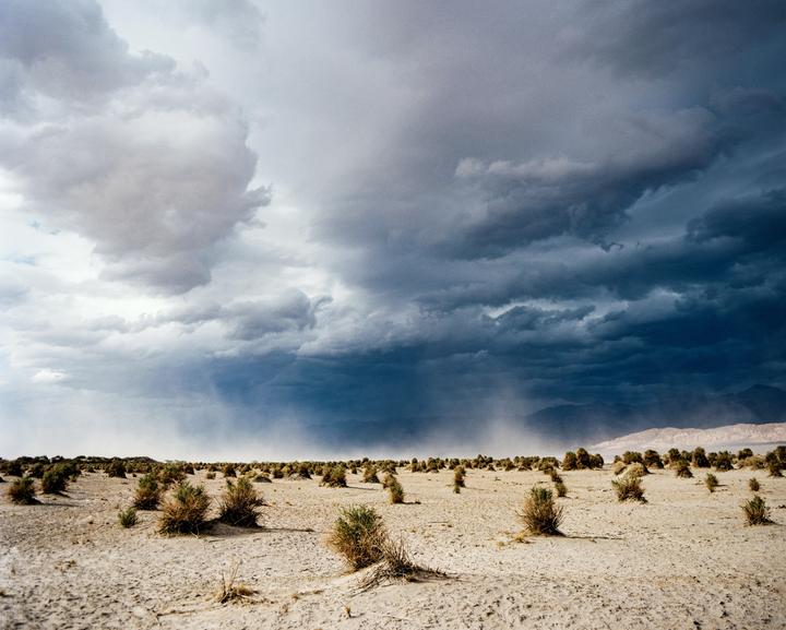 A desert landscape with sparse vegetation is visible under a dramatic sky with dark storm clouds and rain.