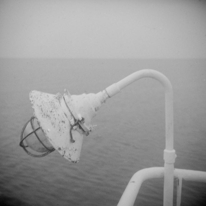 A white lamp with a cage is attached to a railing on a boat, with water and a hazy sky in the background.