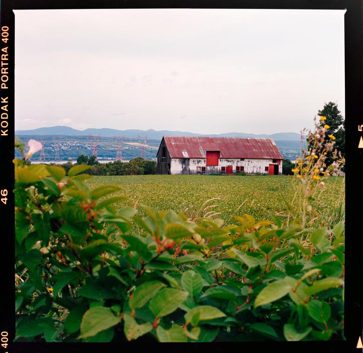 A white barn with a red roof stands in a green field with mountains and a body of water in the distance.