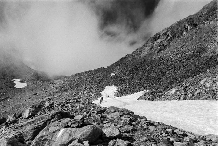 A group of people walk along a snowy path through a rocky mountain landscape under a cloudy sky.