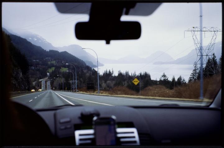 A view from inside a car shows a highway winding through mountains alongside a body of water.
