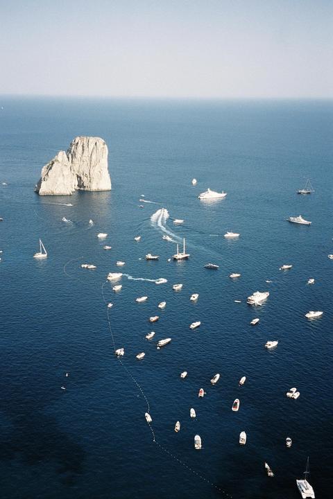 A large rock formation rises from the sea, surrounded by numerous boats and yachts.