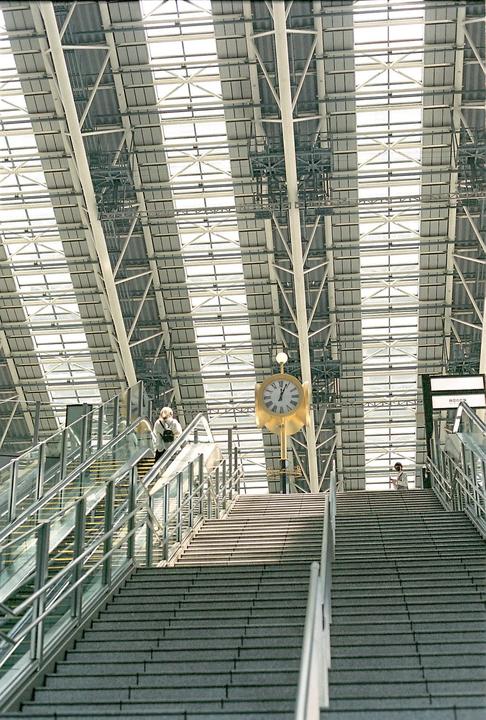 A large golden clock hangs above a wide staircase and escalator in a modern building with a glass and metal roof.