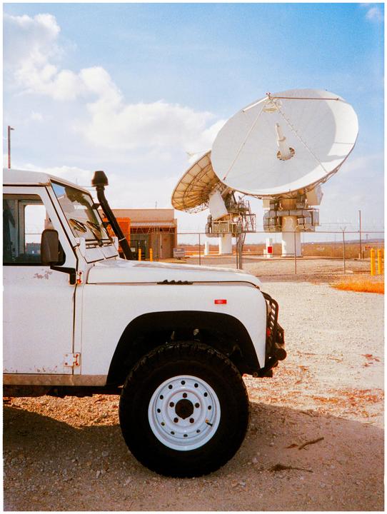 A white off-road vehicle is parked on a gravel surface with large satellite dishes in the background under a blue sky.