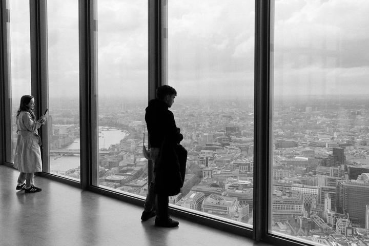 A man and a woman look out of a large window at a city.