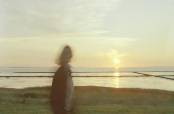 A blurry figure stands in a grassy field overlooking a body of water with the sun setting on the horizon.